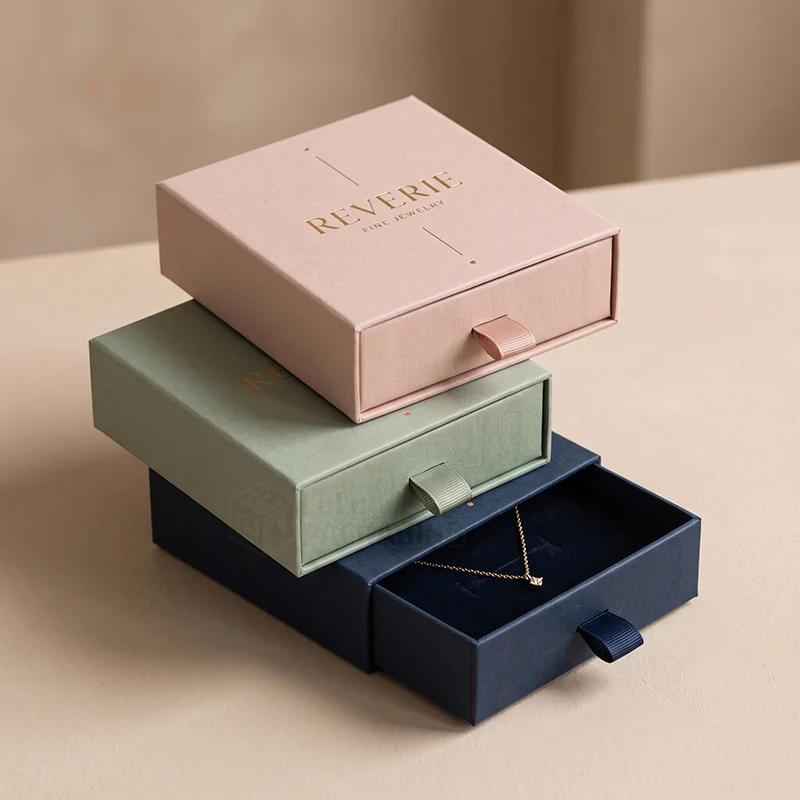 Stack of pink, sage and navy drawer jewellery boxes with ribbon pulls, one open with a necklace
