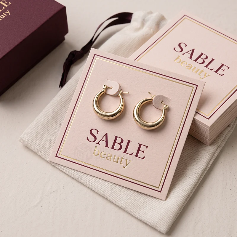 Pink earring card with a gold foil border holding chunky gold hoops on a cotton pouch