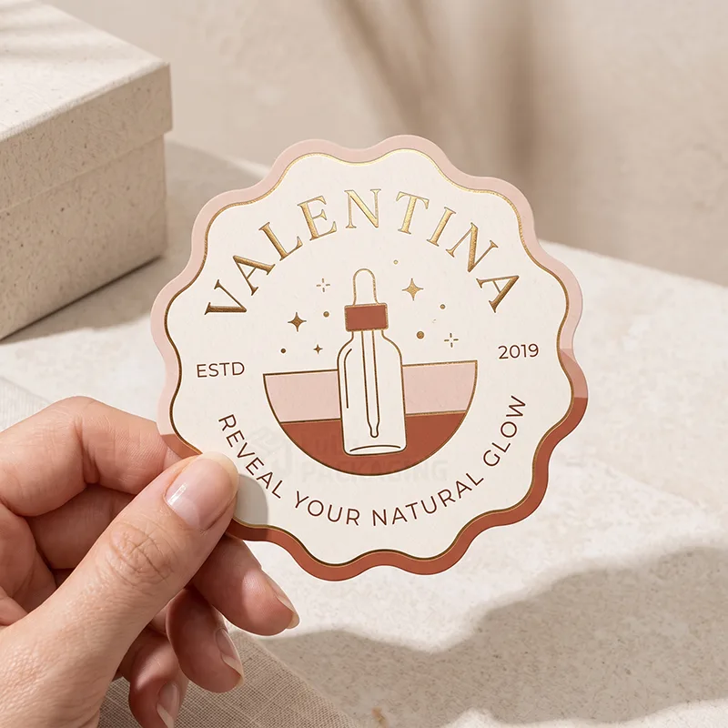 Hand holding a scalloped cream logo sticker with gold foil text and a dropper bottle design