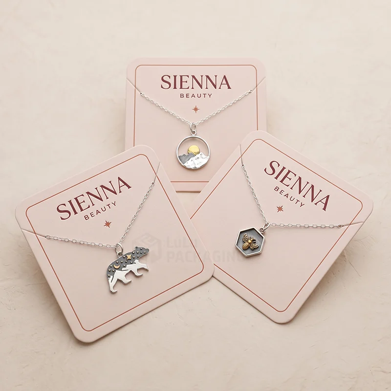 Three pink necklace cards with a printed logo holding silver bear, mountain and bee pendants