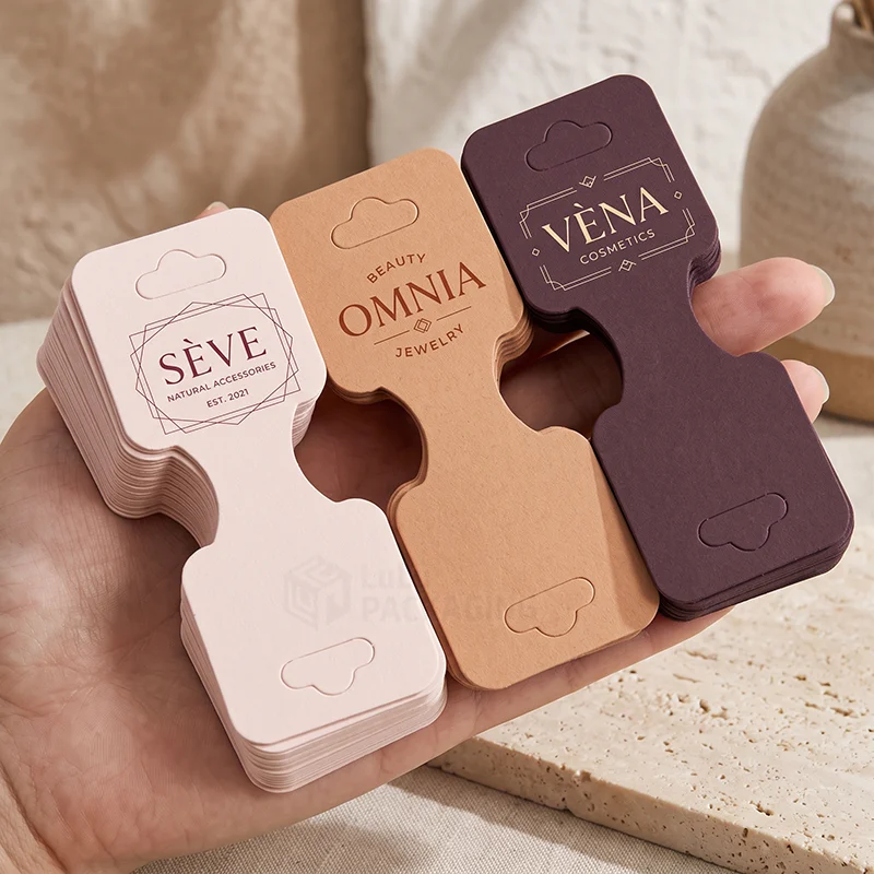 Hand holding stacks of fold-over jewellery display cards in pink, kraft and plum with hang slots