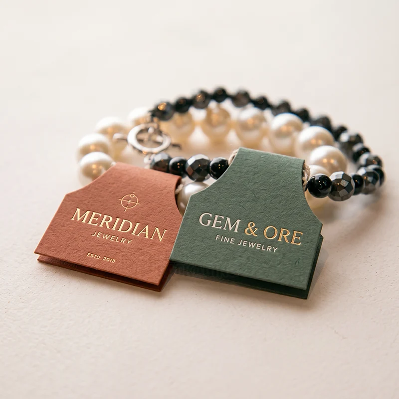 Rust and green fold-over jewellery hang tags with gold foil logos on a pearl bracelet