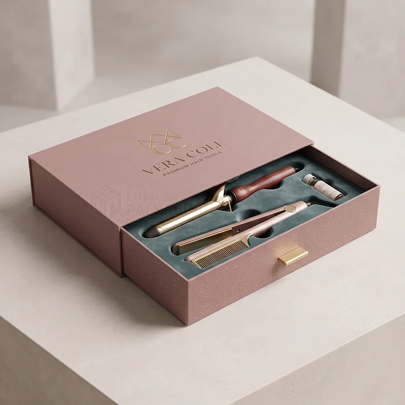 Blush rigid drawer box with gold foil logo holding a curling iron, straightener and comb in a velvet insert