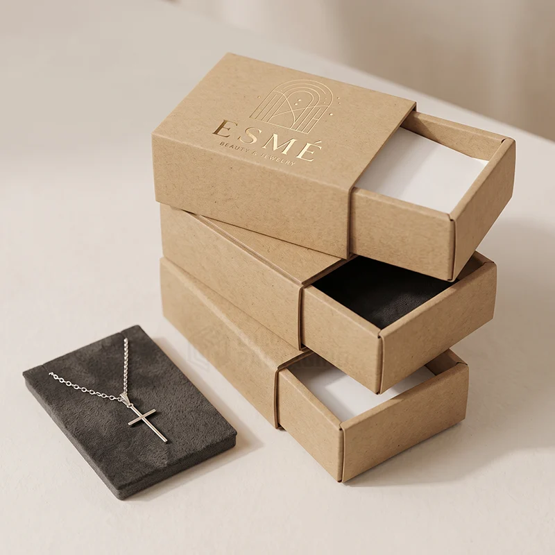 Stack of kraft drawer jewellery boxes with a gold foil logo beside a cross necklace