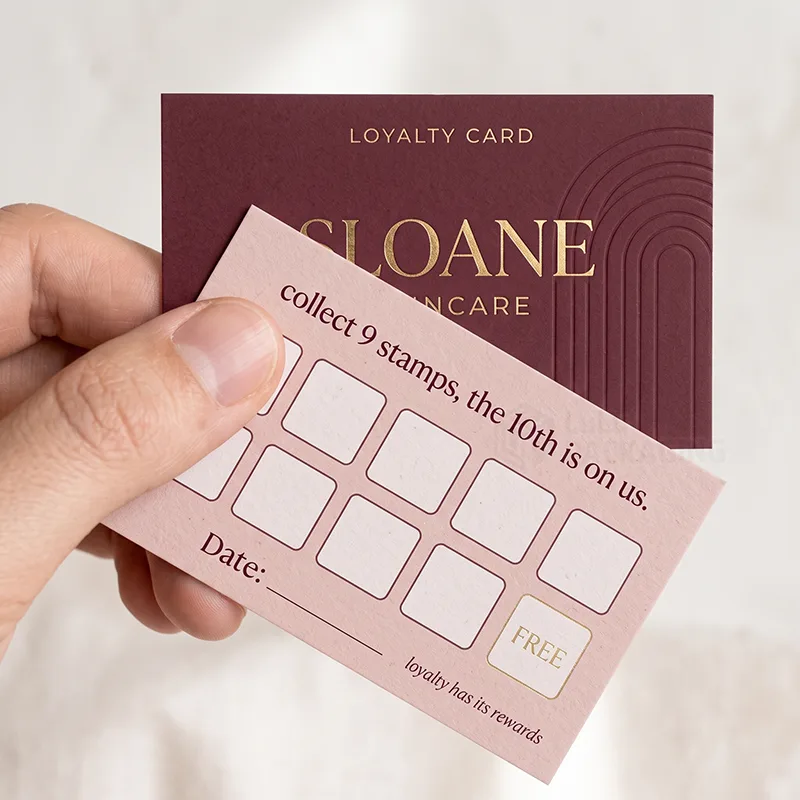 Hand holding a blush stamp grid loyalty card with gold foil free box over a plum foil logo card