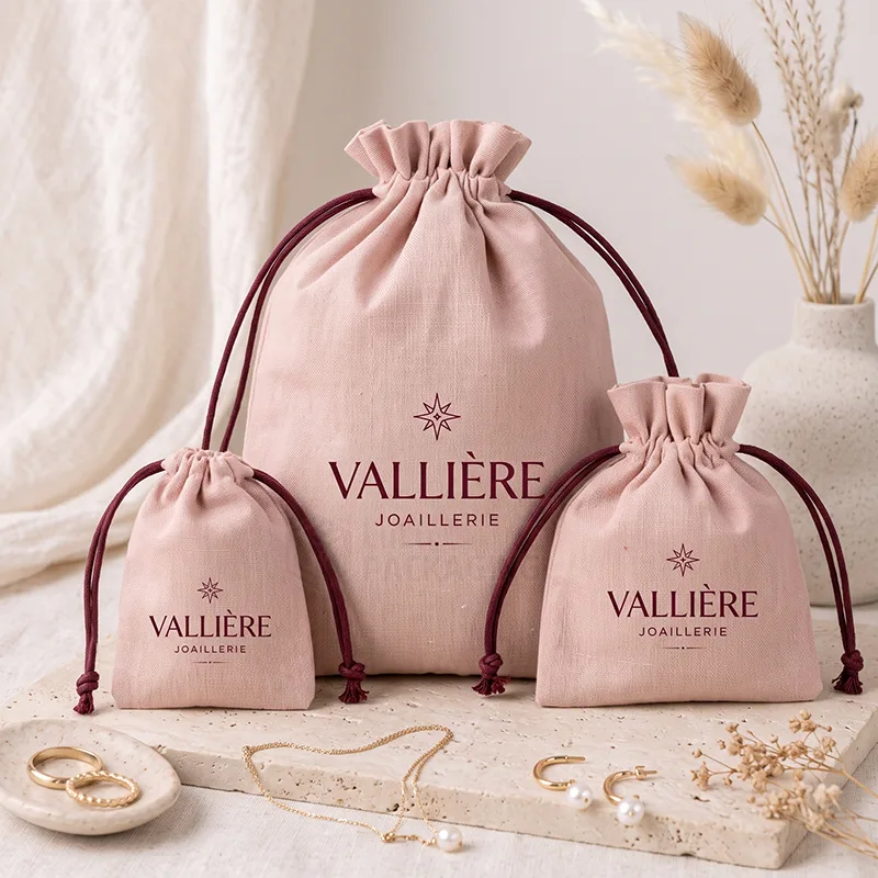 Three blush pink cotton drawstring pouches in different sizes with burgundy cords and a printed logo
