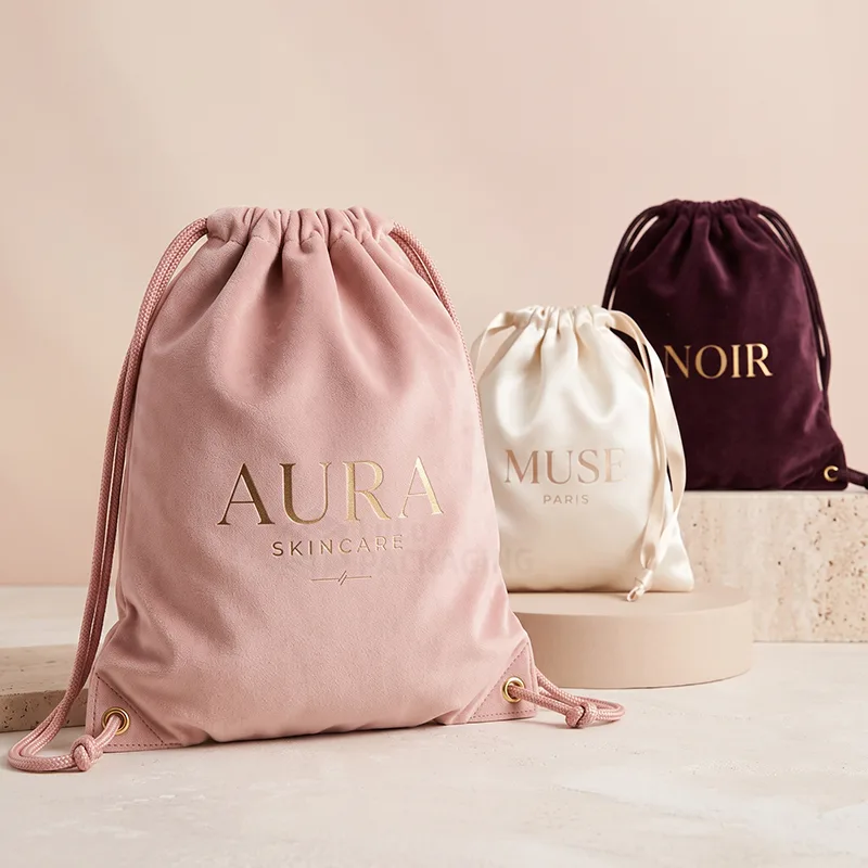 Pink velvet drawstring bag with a gold foil logo beside cream satin and plum velvet bags