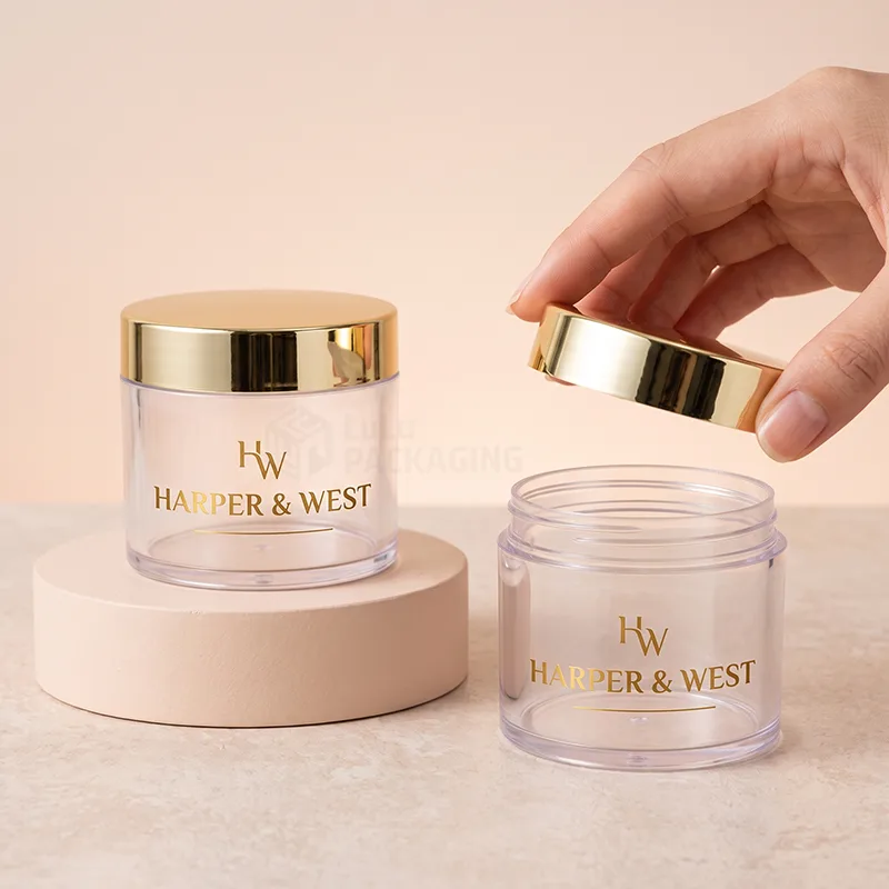 Clear plastic cosmetic jars with shiny gold lids, one lid lifted by hand