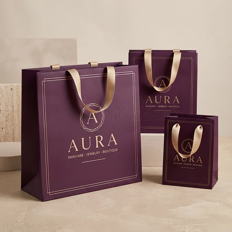 Three purple paper retail bags in different sizes with gold ribbon handles and a gold foil logo