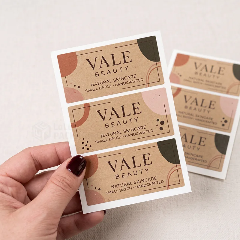 Hand holding a sheet of rectangular kraft labels with terracotta and olive shapes