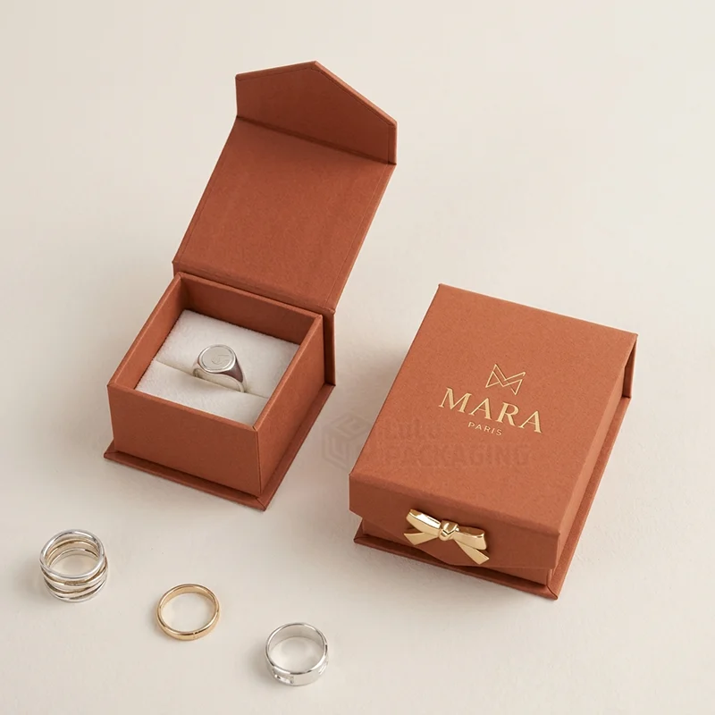 Rust magnetic ring box with a gold foil logo and bow, one open with a signet ring