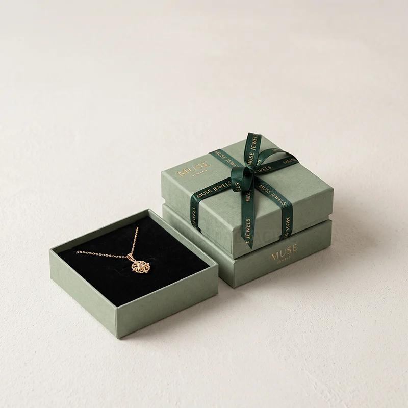 Sage pendant box with a printed green ribbon beside an open box with a gold pendant on black