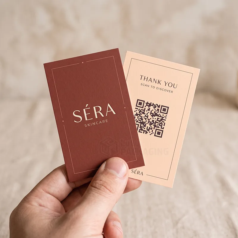 Hand holding a terracotta logo card and a peach thank you card with a scan to discover QR code