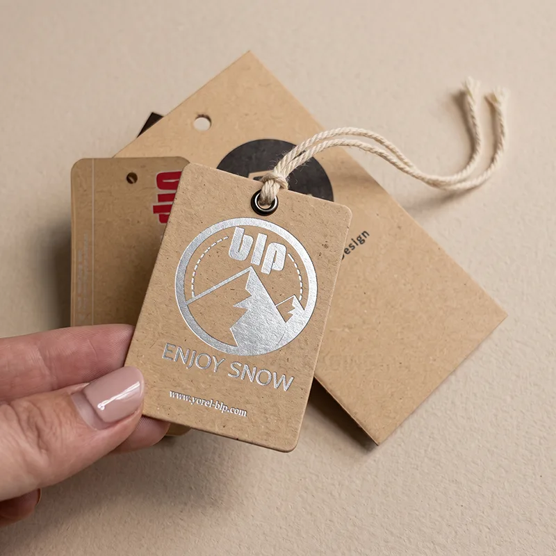 Hand holding a kraft tag with a silver foil mountain logo and Enjoy Snow text, with more kraft tags behind