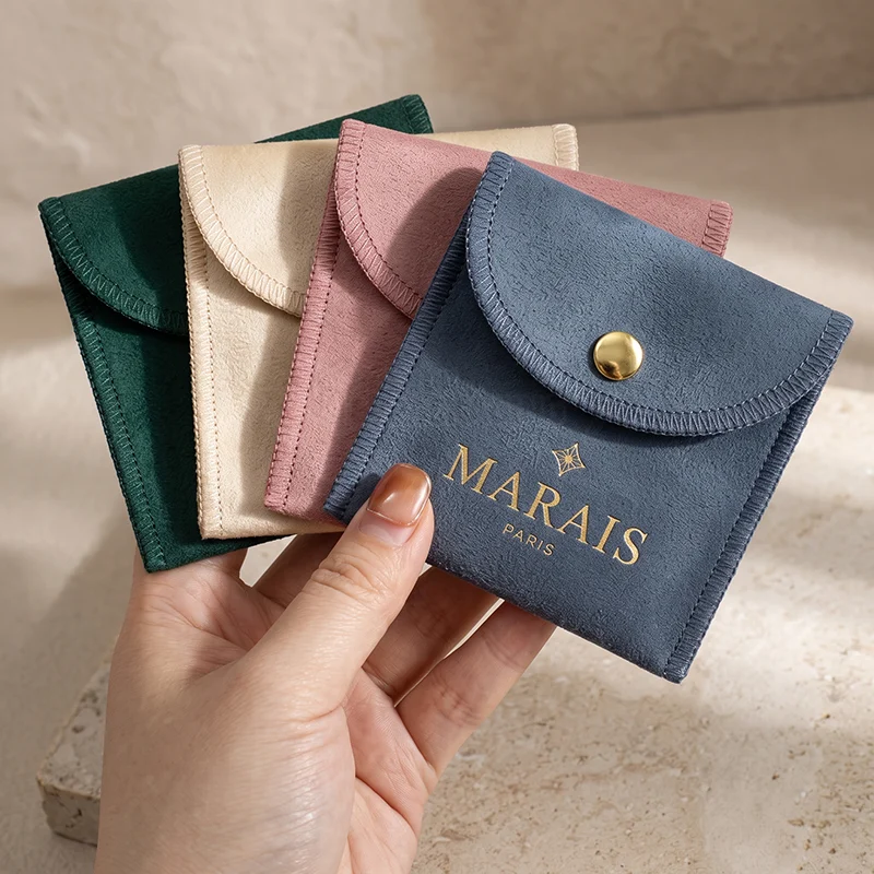 Hand holding velvet envelope pouches in green, cream, pink and blue with a gold snap button