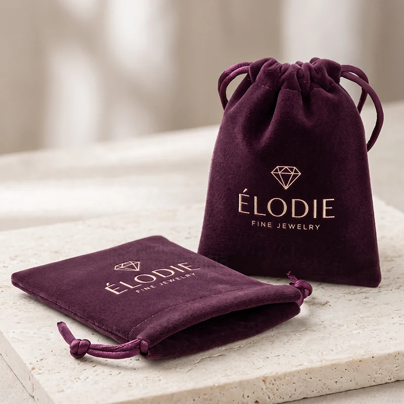 Two plum velvet drawstring pouches with a rose gold foil logo on a stone slab